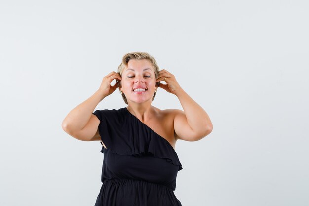 Hermosa mujer posando como usando auriculares en blusa negra y mirando complacido