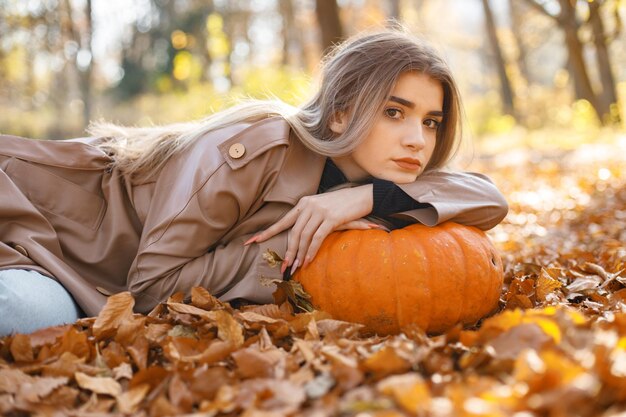 Hermosa mujer posando para una foto en el parque de otoño. Niña acostada sobre hojas amarillas cerca de calabaza. Mujer rubia con abrigo beige.