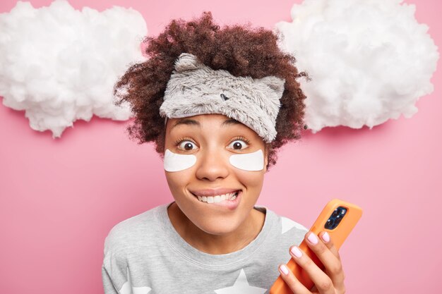 Hermosa mujer positiva muerde los labios mira con alegría a la cámara utiliza el teléfono móvil para navegar por las redes sociales vestida con ropa de dormir aislada sobre una pared rosa. Buenos dias.