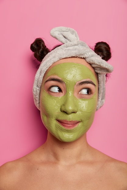 Una hermosa mujer de raza mixta pensativa se preocupa por la piel y el cutis, se aplica una nutritiva mascarilla verde en la cara, usa una diadema, dos moños, se para los hombros desnudos, concentrada a un lado. Procedimientos de spa