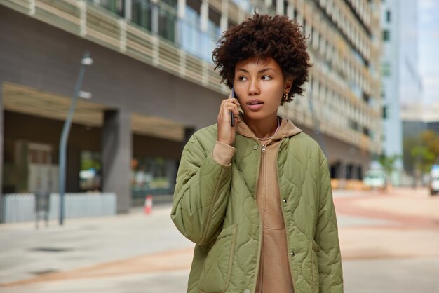 Hermosa mujer reflexiva tiene conversación telefónica mientras camina al aire libre en lugares urbanos posa contra edificios de la ciudad usa chaqueta usa gadget moderno. Concepto de estilo de vida y tecnología de personas