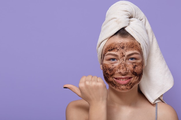 Hermosa mujer relajada y sonriente señala con el dedo pulgar sobre el espacio libre, aplica una máscara de chocolate natural en la cara