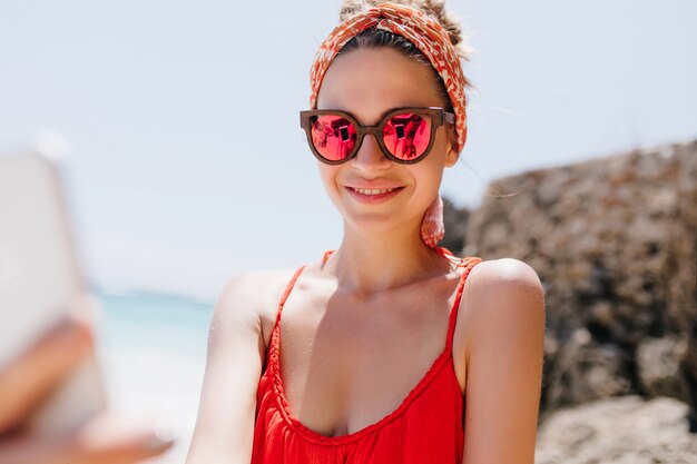 Hermosa mujer riendo haciendo selfie en isla exótica. Foto al aire libre de una chica increíble con gafas de sol tomando una foto de sí misma.