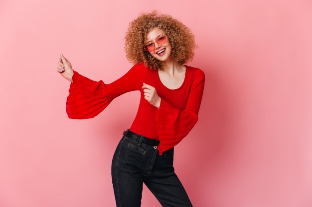 Hermosa mujer rizada bailando en el espacio rosa. Retrato de rubia con gafas rosas con blusa roja y jeans.