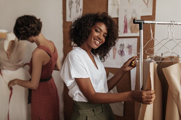 Hermosa mujer rizada de piel oscura en blusa blanca sonríe, mira al frente y trabaja como diseñadora de moda con su amiga