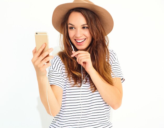 Hermosa mujer en ropa de verano casual hipster sin maquillaje en sombrero marrón escuchando música en el teléfono con auriculares aislados en blanco