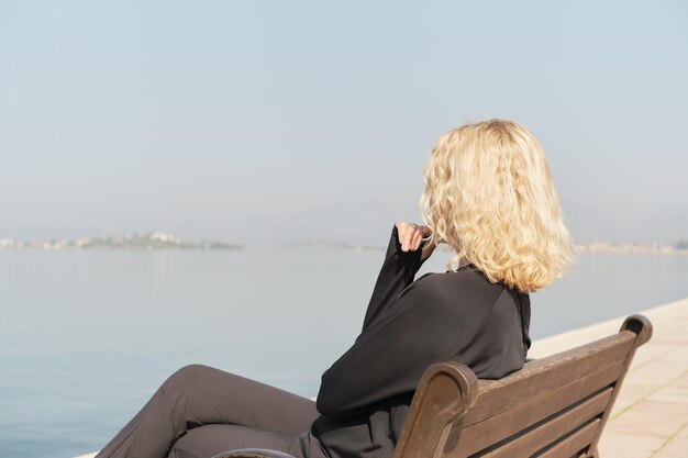 Hermosa mujer rubia adulta se sienta en un banco y mira el mar y el cielo azul silueta de cerca de un espacio de mujer para la idea de texto para el fondo