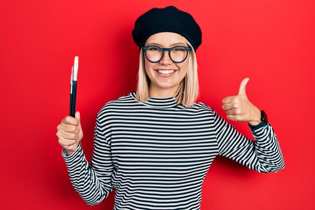 Hermosa mujer rubia con aspecto francés con boina sosteniendo pinceles de pintor sonriendo feliz y positiva, pulgar arriba haciendo excelente y signo de aprobación