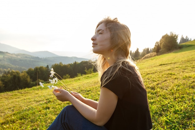 Foto gratuita hermosa mujer rubia disfrutando de vistas a las montañas, sosteniendo camomiles