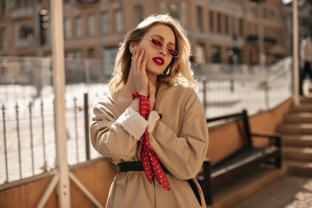 Hermosa mujer rubia elegante con labios rojos en gafas de sol de colores brillantes y gabardina beige toca suavemente la cara y posa afuera