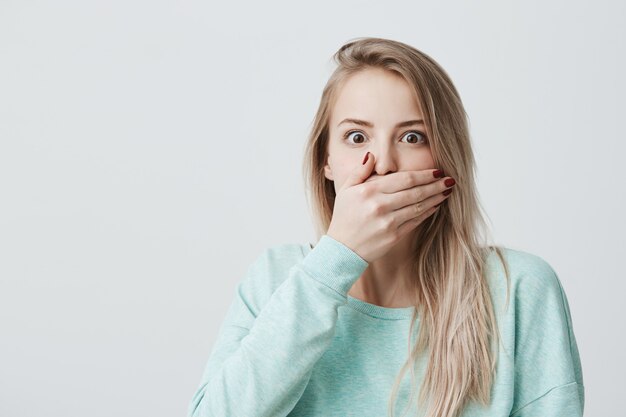 Hermosa mujer rubia emocionada cubre la boca con asombro, sus ojos se reventaron