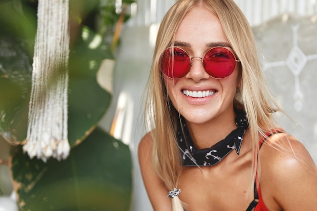 Hermosa mujer rubia con expresión alegre, lleva gafas de sol rojas de moda