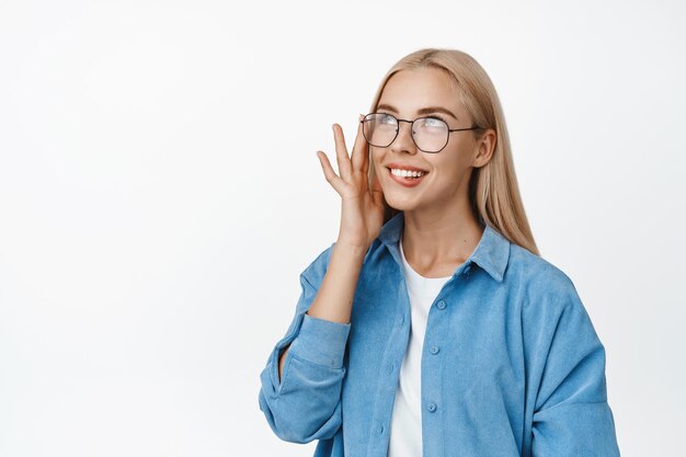 Hermosa mujer rubia con gafas sonriendo y mirando coqueta a la esquina superior izquierda de pie sobre fondo blanco.