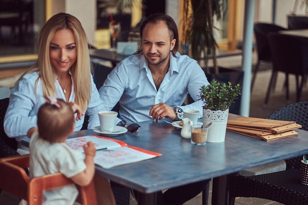Hermosa mujer rubia y hombre guapo disfrutando de la vida familiar con su pequeña hija en un café al aire libre.