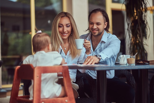 Hermosa mujer rubia y hombre guapo disfrutando de la vida familiar con su pequeña hija en un café al aire libre.