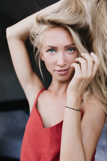 Hermosa mujer rubia con ojos azules relajante por la mañana. Foto interior de adorable dama blanca jugando con su cabello rubio.