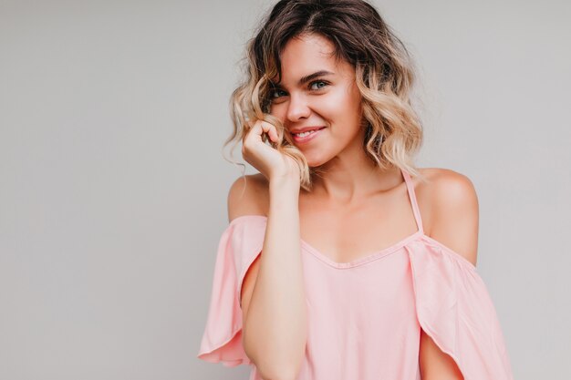 Hermosa mujer rubia posando con sonrisa tímida. Filmación en interiores de una chica entusiasta en traje rosa.