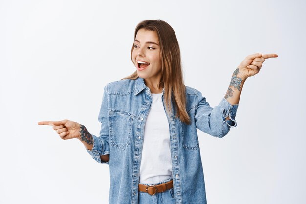 Hermosa mujer rubia con ropa informal apuntando de lado a las variantes de compras y haciendo elecciones indicando y mirando a la izquierda en el espacio de la copia de pie contra el fondo blanco