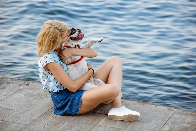 Hermosa mujer rubia sentada, jugando con bulldog francés cerca del mar