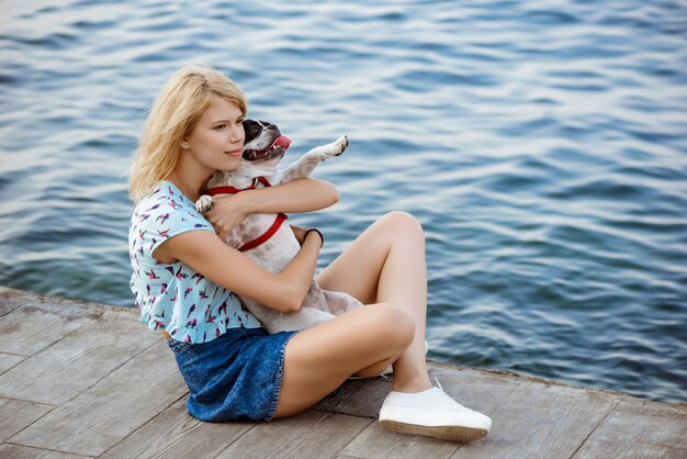 Hermosa mujer rubia sentada, jugando con bulldog francés cerca del mar