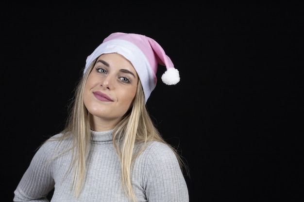 Hermosa mujer rubia con un sombrero rosa de Santa Claus posando sobre una pared negra