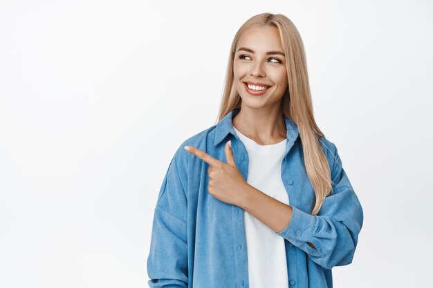 Hermosa mujer rubia sonriendo señalando con el dedo y mirando a la izquierda el logotipo de la empresa que muestra un anuncio de pie sobre fondo blanco.