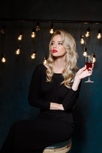 Hermosa mujer rubia en traje de noche posando, sosteniendo una copa de vino