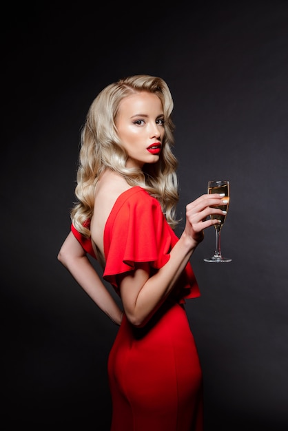 Hermosa mujer rubia en traje de noche posando, sosteniendo cristal de champaña