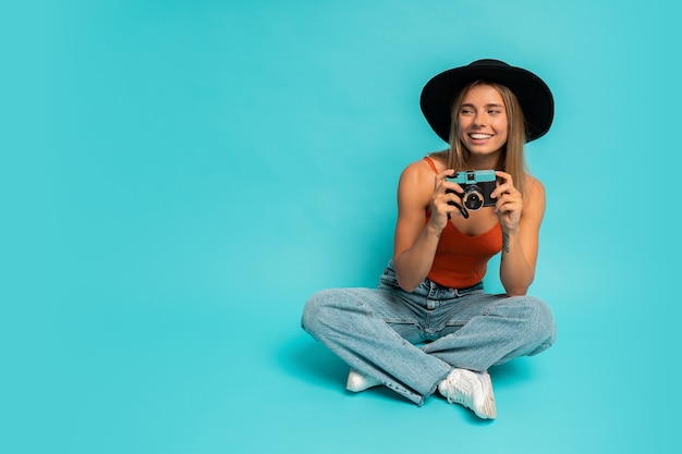 Hermosa mujer rubia en traje de verano con estilo con cámara retro, sentada en el piso en el estudio de la pared azul. Estado de ánimo de vacaciones.