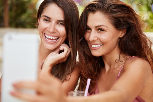 Hermosa mujer satisfecha con apariencia atractiva tiene una sonrisa agradable, sostiene un teléfono inteligente, se sienta cerca de su mejor amiga