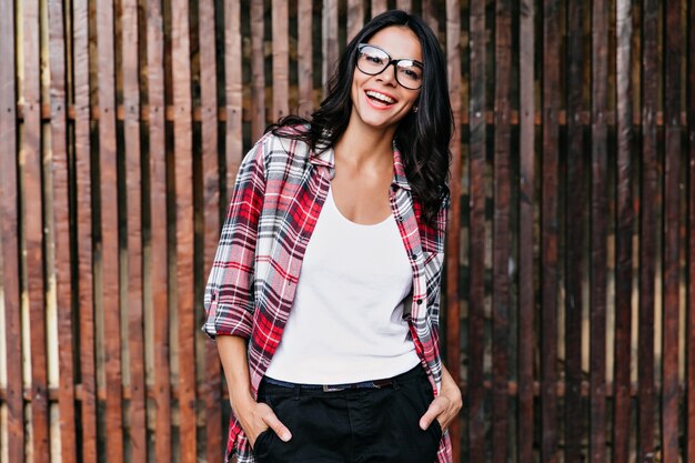 Hermosa mujer segura de pie con las manos en los bolsillos. Retrato al aire libre de una chica morena de ensueño en camiseta blanca aislada en la pared de madera.