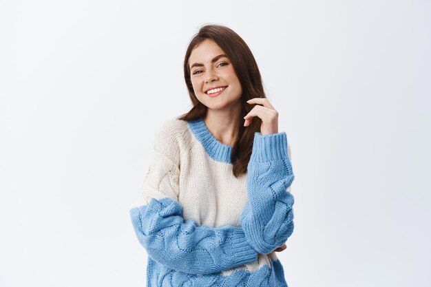 Hermosa mujer segura de sí misma tocando el cabello y sonriendo complacida de tener una idea interesante, de pie seguro de sí mismo en un suéter cálido en blanco