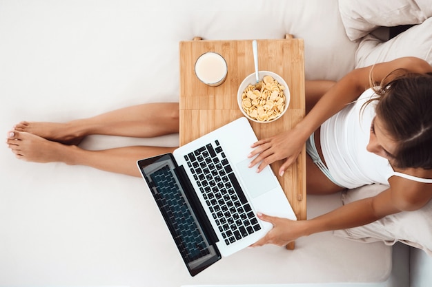 Hermosa mujer sentada en la cama con el portátil, desayunando Disparo desde arriba ..