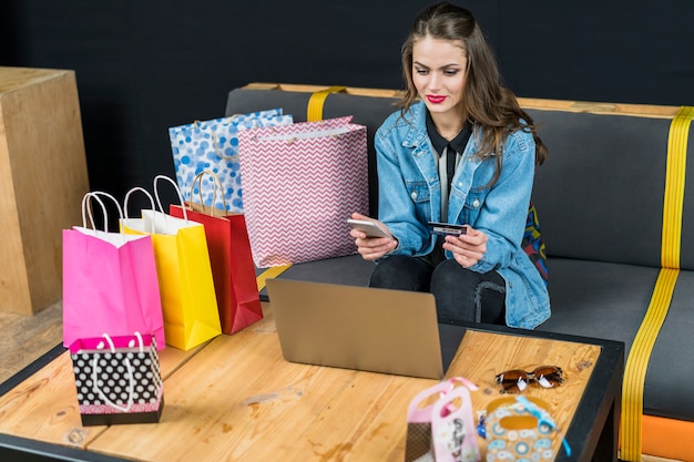 Hermosa mujer sentada en casa con dispositivos electrónicos; Bolsas de compra y tarjeta de crédito en mano.