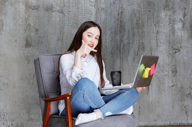 Hermosa mujer sentada en una silla, sosteniendo una taza de café y una computadora portátil. foto de alta calidad