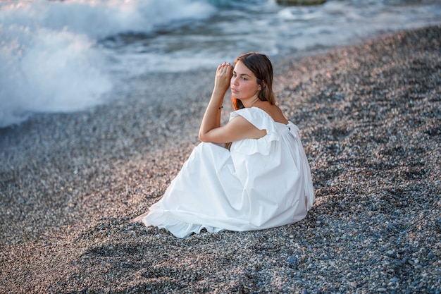 Hermosa mujer sentada sola y pensando en vestido blanco en la orilla del mar y con cara de tristeza.