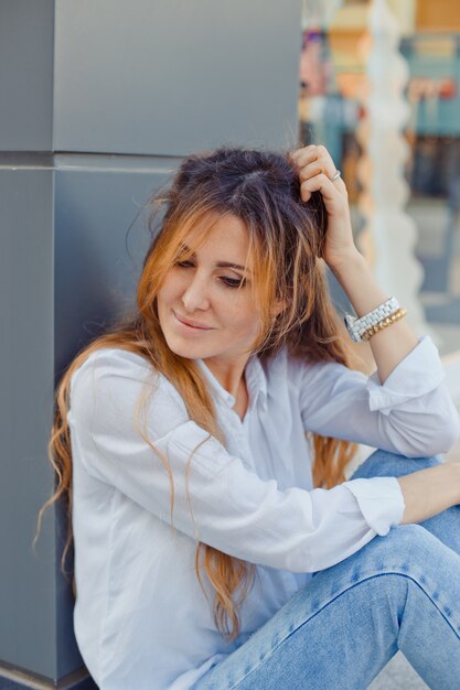 Hermosa mujer sentada en el suelo mirando hacia abajo y sonriendo en la calle durante el día.