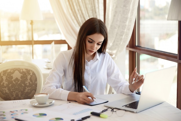 Hermosa mujer seria está escribiendo algo en el cuaderno en su lugar de trabajo