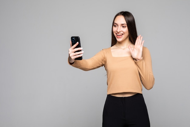 Hermosa mujer sociable con apariencia asiática tomando selfie o hablando por videollamada usando un teléfono celular aislado sobre una pared gris