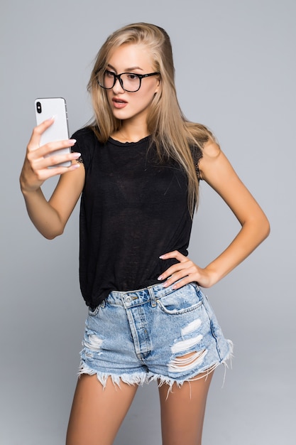 Hermosa mujer sociable tomando selfie o hablando por videollamada con teléfono celular sobre fondo gris