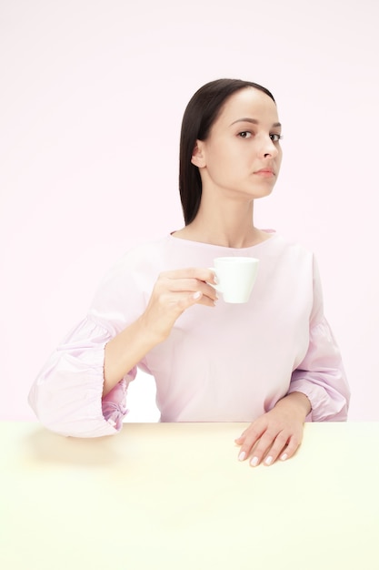 Hermosa mujer solitaria sentada en el estudio rosa y mirando triste sosteniendo la taza de café en la mano. Retrato en tonos de primer plano en estilo minimalista