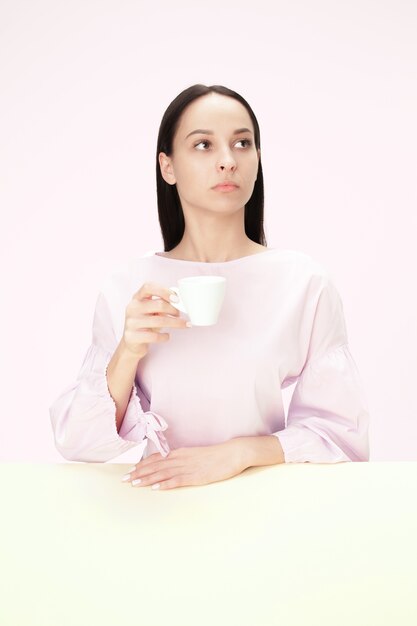 Hermosa mujer solitaria sentada en el estudio rosa y mirando triste sosteniendo la taza de café en la mano.