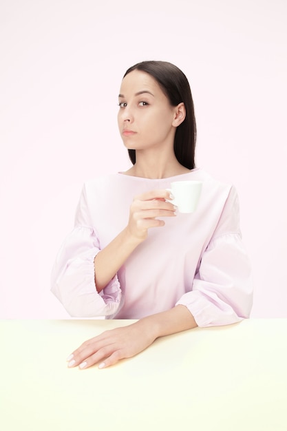 Hermosa mujer solitaria sentada en el estudio rosa y mirando triste sosteniendo la taza de café en la mano.