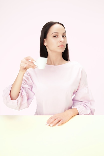 Hermosa mujer solitaria sentada en el estudio rosa y mirando triste sosteniendo la taza de café en la mano.