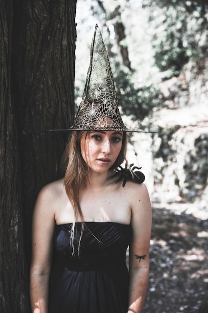Hermosa mujer en sombrero de bruja de pie cerca de árbol y mirando a cámara