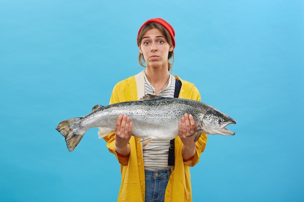 Foto gratuita hermosa mujer con sombrero rojo, impermeable amarillo y sosteniendo un enorme pez en las manos