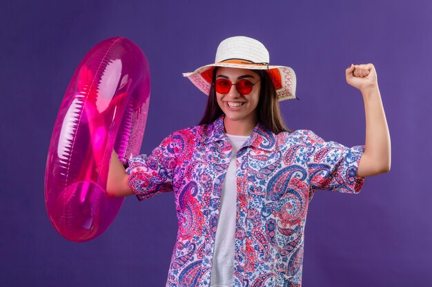 Hermosa mujer con sombrero de verano y gafas de sol rojas con anillo inflable salió regocijándose de su éxito y victoria apretando los puños sonriendo alegremente listo para el concepto de vacaciones de pie