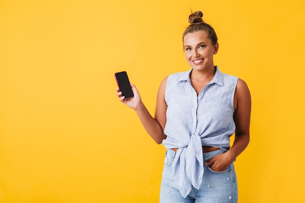 Hermosa mujer sonriente en camisa mirando felizmente a la cámara mientras muestra un nuevo teléfono celular sobre fondo amarillo