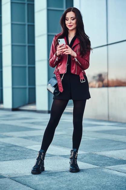 Hermosa mujer sonriente con chaqueta roja está parada al lado de un edificio de vidrio y charlando con alguien por teléfono móvil.