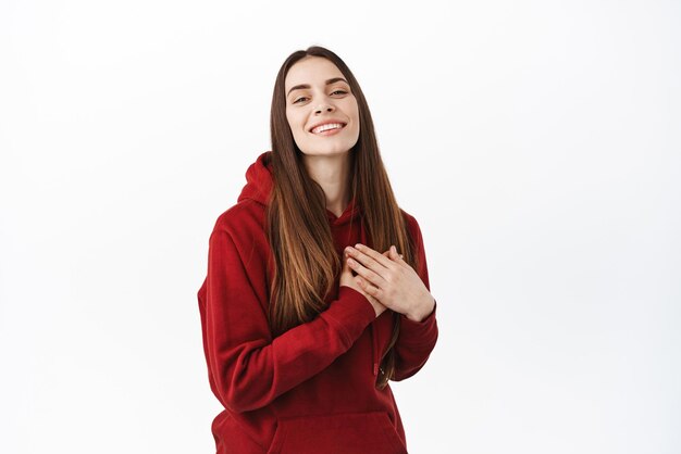 Hermosa mujer sonriente cogida de la mano en el corazón se ve complacida y agradecida recibe un buen regalo aprecia el gesto cálido sintiéndose conmovida y agradecida de pie sobre fondo blanco